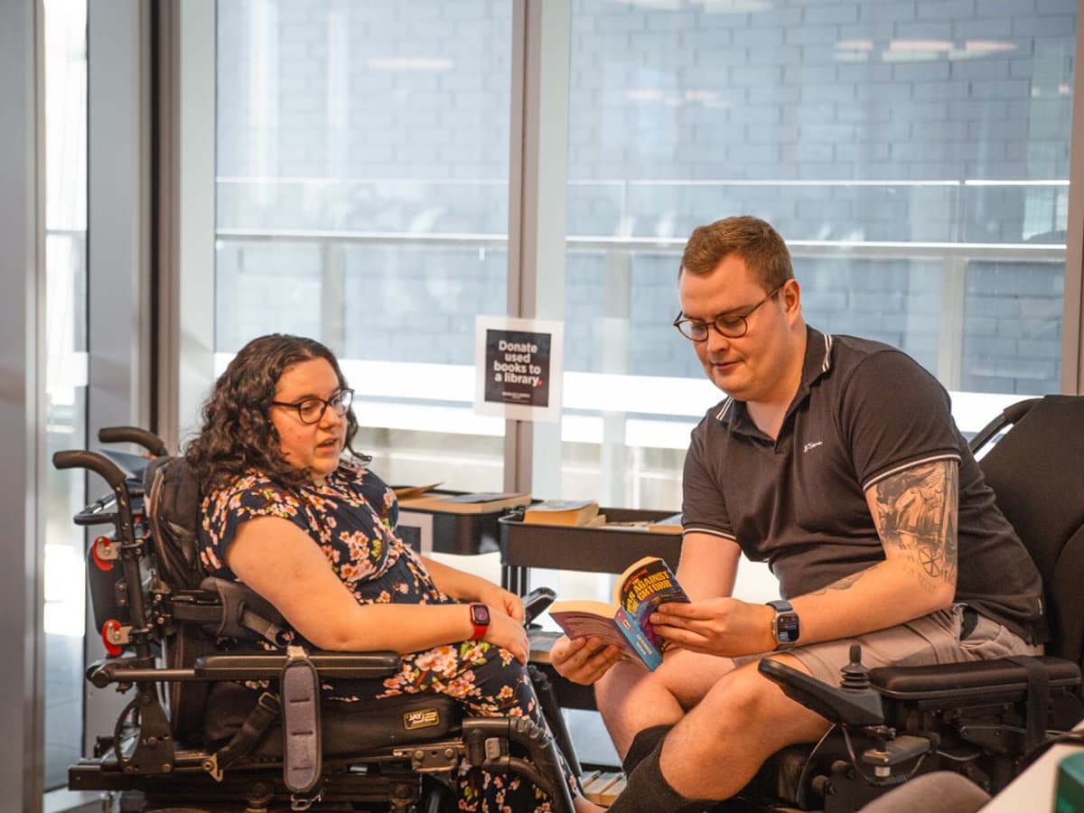 Support worker and participant engaged in relaxed conversation and reading together in a comfortable lounge