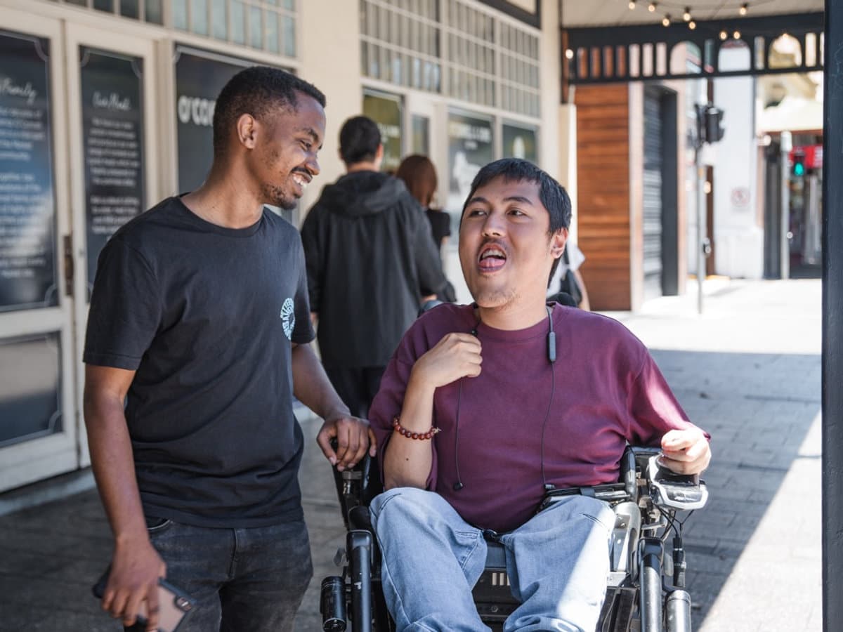 Cheerful Australian participant in an electric wheelchair smiling with a friendly support worker during a community outing